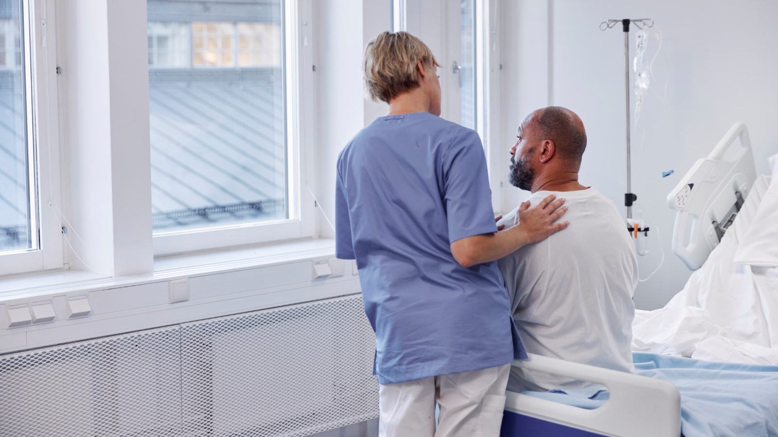 En sjuksköterska klappar en patient på ryggen, de tittar ut genom ett sjukhusfönster. 